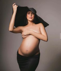 a pregnant woman in a black dress and hat poses for the camera with her hands on her head