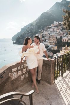 a man and woman standing next to each other on top of a balcony near the ocean