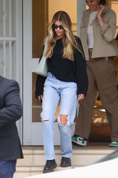 a woman in ripped jeans and black sweater walking out of a building with her hand on her hip