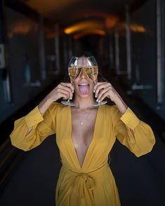 a woman in a yellow dress holding two wine glasses up to her face with both hands