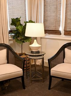 two chairs and a table with a lamp on it in a living room next to windows