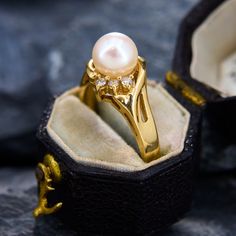 a gold ring with a pearl and diamonds in it sitting inside a box on the ground