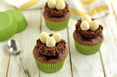 four cupcakes with chocolate frosting and banana toppings on top, sitting next to a spoon