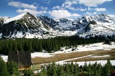 the mountains are covered in snow and pine trees, with a small cabin on the far side