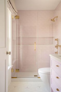 a white toilet sitting next to a walk in shower under a bathroom window with gold fixtures