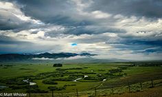 an aerial view of a green valley with mountains in the distance and clouds above it