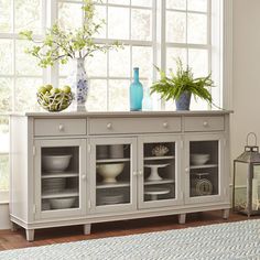 a white sideboard with glass doors and drawers in front of a blue vase on the window sill