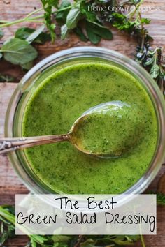 a spoon in a jar filled with green liquid surrounded by fresh herbs and greenery