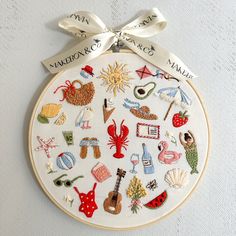 a white embroidered wall hanging with various items on it and a ribbon tied around the hoop