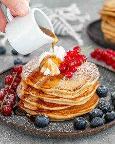 a person pouring syrup onto a stack of pancakes on a plate with berries and blueberries