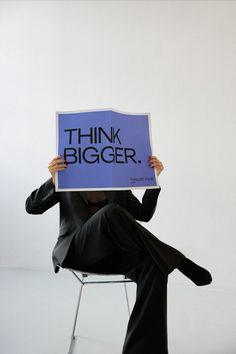 a person sitting in a chair holding up a sign that says think bigger on it