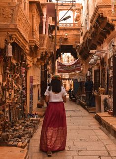 a woman in a long red skirt walking down an alley way with shops on both sides