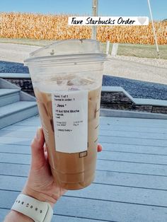 someone holding up a cup of iced coffee in front of a corn field with the words fave starbucks order written on it