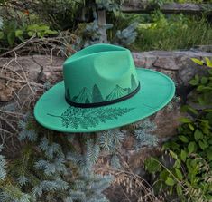 This hand burnt wide brim fedora makes a perfect addition to any nature lover's  wardrobe. The Kelly green material burns a gorgeous shade of emerald green allowing the design to stand out. I burnt pine trees in front of a mountain scene around the entire crown of the hat. Each mountain is intricately burned by hand with a pyrography machine using various tips to achieve depth and a rock-like texture.  On the right side brim of the hat I burnt a realistic fern. For a finishing touch, I burnt dis Custom Brimmed Fedora For Outdoor, Custom Handmade Hats For Outdoors, Artisan Green Hat With Flat Brim, Bohemian Felt Hat With Curved Brim For Outdoor, Custom Green Hat With Curved Brim, Handmade Green Bohemian Fedora, Artisan Style Fedora With Flat Brim For Outdoor, Green Fedora With Flat Brim For Outdoor, Artisan Green Hat With Curved Brim