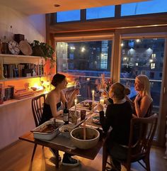 three women sitting at a table with food and drinks in front of them, looking out the window