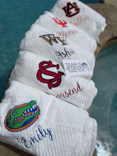 four white towels with embroidered logos on them sitting next to a body of water in the background