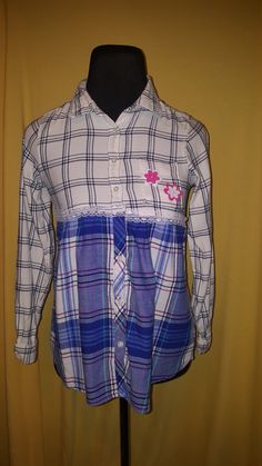 This upcycled flannel shirt features a white/dark blue plaid top, complimented with a blue/white plaid shot with green and pink, and lace trim on the waist and collar.  Pink and white felt flowers finish it off. White Cotton Flannel Shirt For Spring, Preppy Plaid Cotton Top, Spring Gingham Flannel Cotton Shirt, Blue Cotton Flannel Shirt For Spring, Cotton Button-up Shirt With Lace Trim, Cotton Lace Trim Button-up Shirt, Plaid Flannel Shirt For Spring, Spring Plaid Flannel Shirt, Casual Button-up Shirt With Lace Trim