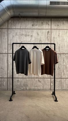 three t - shirts are hanging on a rack in front of a concrete wall and metal pipe