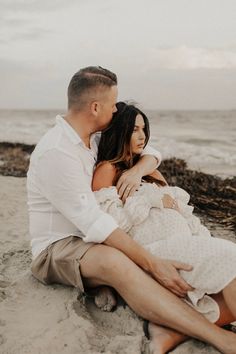 a man and woman are sitting on the sand by the ocean with their arms around each other