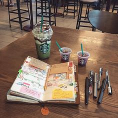 an open book sitting on top of a wooden table next to cups of coffee and writing utensils