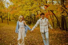 a man and woman holding hands while walking through the woods with leaves on the ground