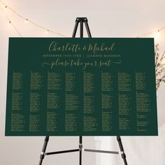 a green and gold wedding seating chart on a easel with lights in the background