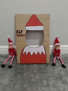 two elfs sitting in front of a cardboard box with the door cut out to look like an elf