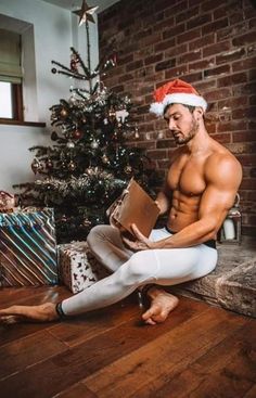 a shirtless man sitting on the floor next to a christmas tree holding a box