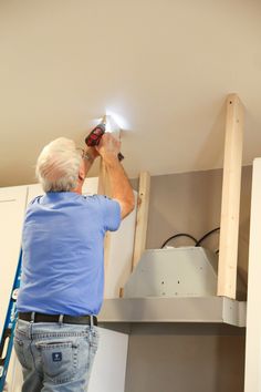 a man working on a light fixture in a room with white cabinets and ladders