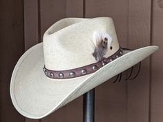 Chet starts with a Mexican soft palm leaf hat body that has a 3 3/4" brim and 5 1/4" crown. The band is simple with distressed brown leather and antique nickel color spots. The back of the band has a deer antler bead for accent. A neutral feather combo completes the hat. Note: Please allow 3-4 weeks. Adjustable Distressed Brown Country Hat, Adjustable High Crown Straw Hat In Natural Color, Distressed Brown Adjustable Hat With Flat Brim, Adjustable Straw Hat With Flat Crown For Country Events, Distressed Brown Adjustable Short Brim Hat, Distressed Brown Adjustable Flat Brim Hat, Distressed Brown Adjustable Hat For Western-themed Events, Rustic Adjustable Hat Bands In Distressed Brown, Adjustable Distressed Brown Hats For Rodeo