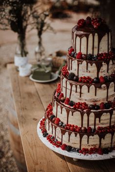 a three tiered cake with chocolate icing and berries on the top is sitting on a wooden table