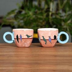 two coffee mugs with birds painted on them sitting on a wooden table next to a potted plant