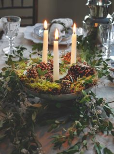 three candles are lit in a bowl with moss and pine cones on the centerpiece