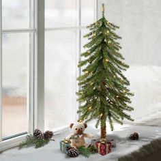 a teddy bear sitting next to a small christmas tree in front of a large window