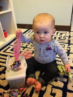 a toddler sitting on the floor holding an object in her hands and looking at the camera