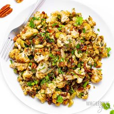 a white plate topped with cauliflower and nuts next to a fork on top of a table