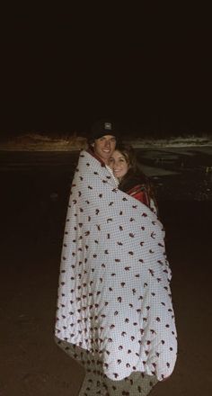 a man and woman wrapped up in a blanket on the beach at night with their arms around each other