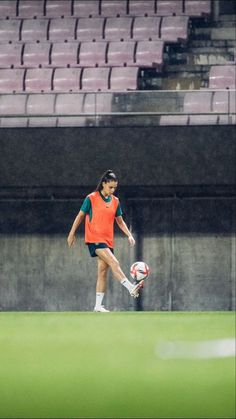 a woman kicking a soccer ball on top of a lush green field in front of an empty stadium