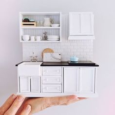 a hand holding a model of a kitchen with white cabinets and black counter tops,