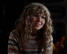 a woman with glasses is smiling and looking at the camera while sitting in a dark room