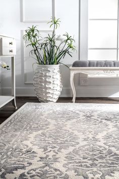 a white vase with some green plants in it next to a rug on the floor