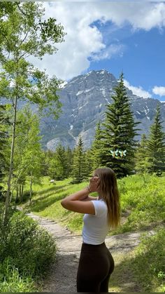 a woman standing on a dirt path in the woods looking up at mountains and trees