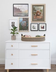 there is a white dresser with pictures on the wall and a basket in front of it