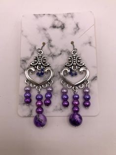 2 Inch drop-earrings made with hypoallergenic materials.   Beads are attached to a silver plated heart charm consisting of a purple marble bead and three smaller glass beads in three different shades of purple/lavender in the center.  Left/Right sides contain the same smaller glass beads in the three different shades of purple/lavender as the center.  The middle of the heart charm is accented with four tiny glass beads as an accent.  These beads are a darker shade of purple. Includes soft ear backings. Materials are environmental, lead free and non-toxic. Light weight, smooth surface, and a touch of sheen to add elegance. The earrings are attached to a marble cardboard earring holder and are shipped in a sealed plastic cover, and placed inside a mesh drawstring "thank you" pouch as shown. Purple Metal Chandelier Earrings For Gifts, Purple Metal Chandelier Earrings As Gift, Hypoallergenic Purple Beaded Earrings, Purple Teardrop Metal Earrings, Purple Dangle Heart Earrings For Pierced Ears, Purple Drop Chandelier Earrings As Gift, Purple Metal Dangle Earrings, Nickel-free Purple Beaded Earrings, Nickel-free Purple Beaded Earrings With Round Beads