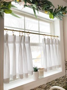 a kitchen window with white curtains and green plants hanging from the windowsill, next to a potted plant