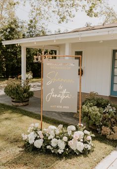 a wedding sign in front of a white house with flowers on the lawn and lights hanging from the roof