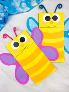 two paper crafts with eyes and antennae on top of a marble surface, one is yellow and the other is blue