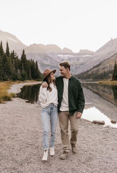 a man and woman are walking by the water in their outfits, with mountains in the background