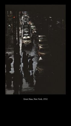 people are standing on the sidewalk in the rain near parked cars and street lights at night