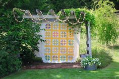 a quilt hanging on a wooden structure in the grass with flowers and greenery around it